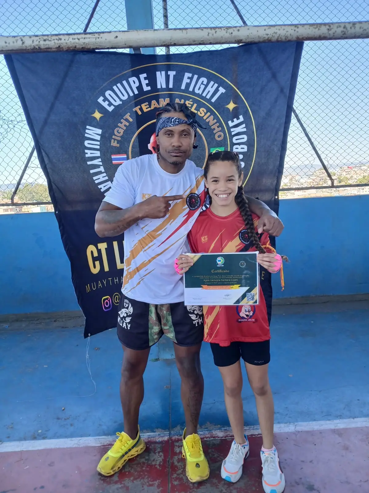 Aluna da NT Fight com o certificado de graduação nas mãos, ao lado do Prof. Nélsinho, diante da bandeira da equipe
