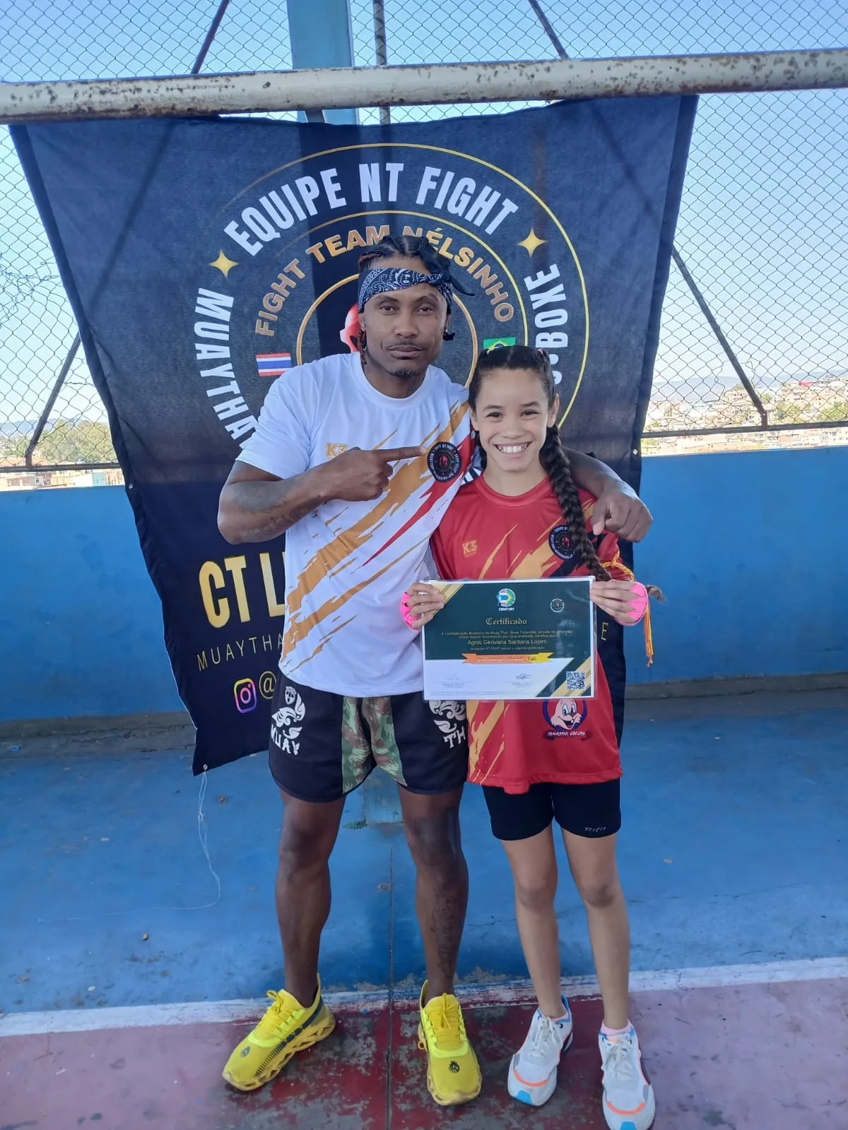 Aluna da NT Fight com o certificado de graduação ao lado do Prof. Nélsinho, diante da bandeira da equipe
