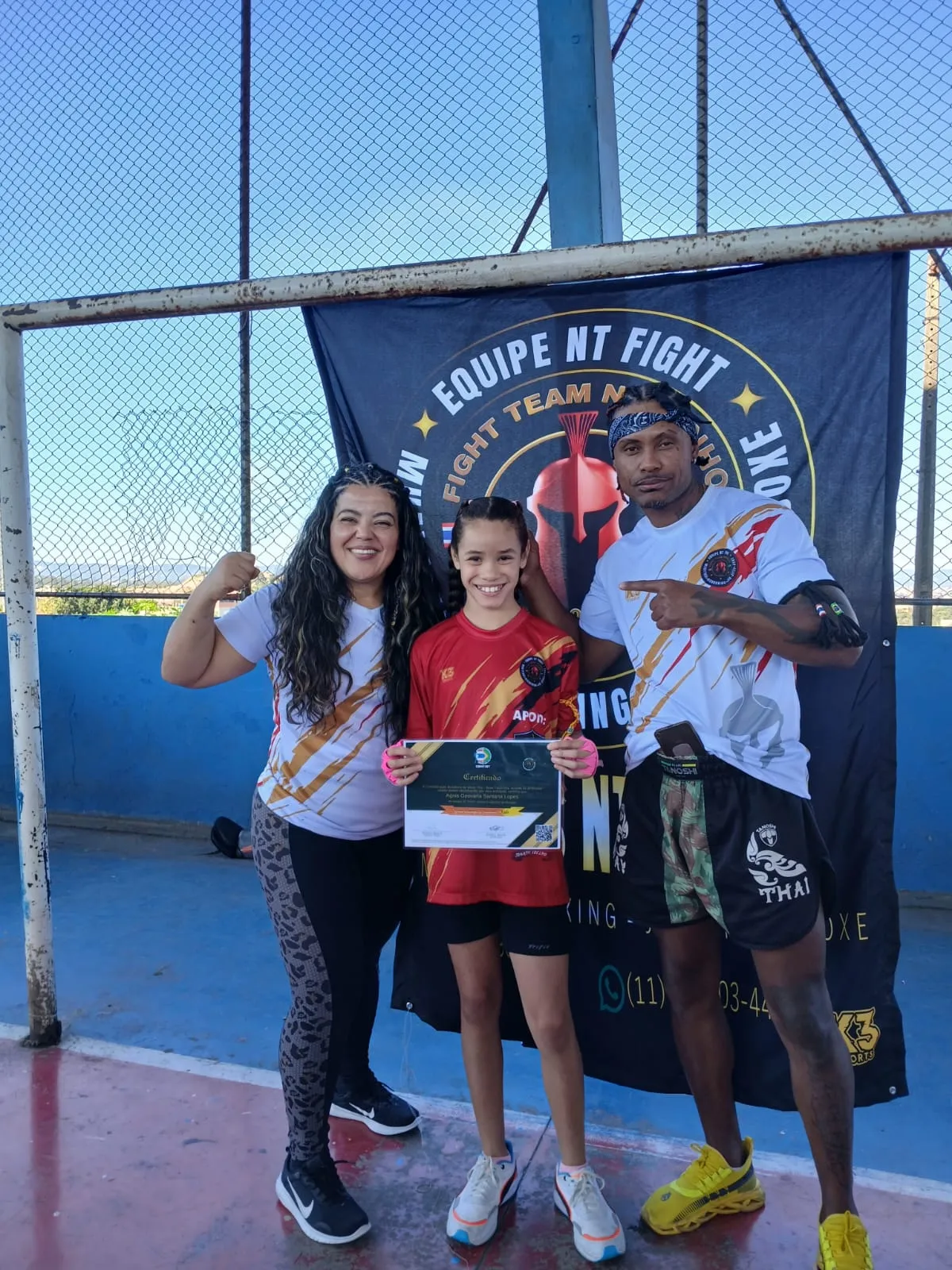 Aluna da turma kids acompanhada da mãe e do Prof. Nélsinho diante da bandeira da Equipe NT Fight