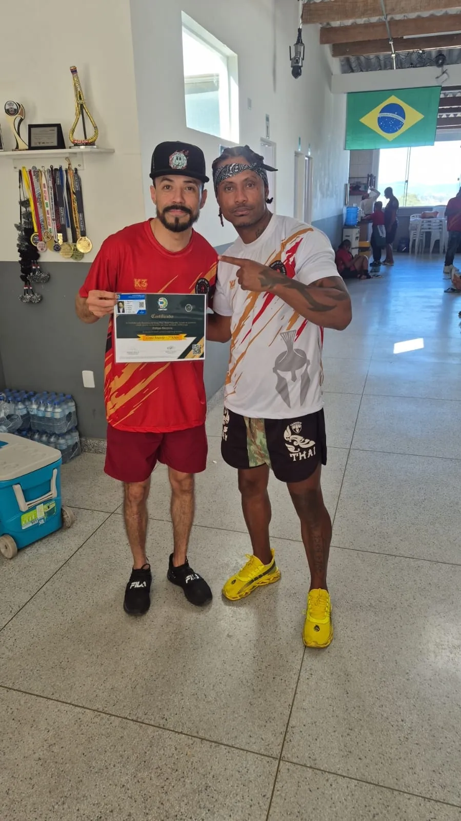 Aluno com o certificado ao lado do Prof. Nélsinho no CT da equipe, sob a prateleira de troféus