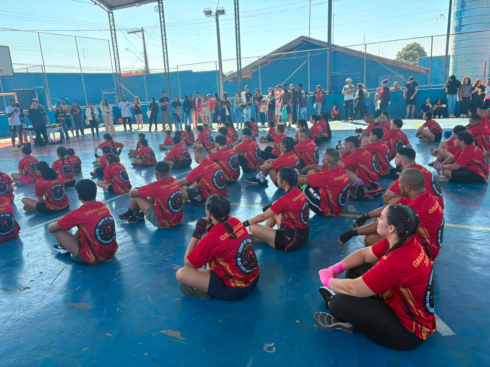 Alunos de camisa da Equipe NT Fight sentados na quadra durante a cerimônia, com as famílias assistindo ao fundo