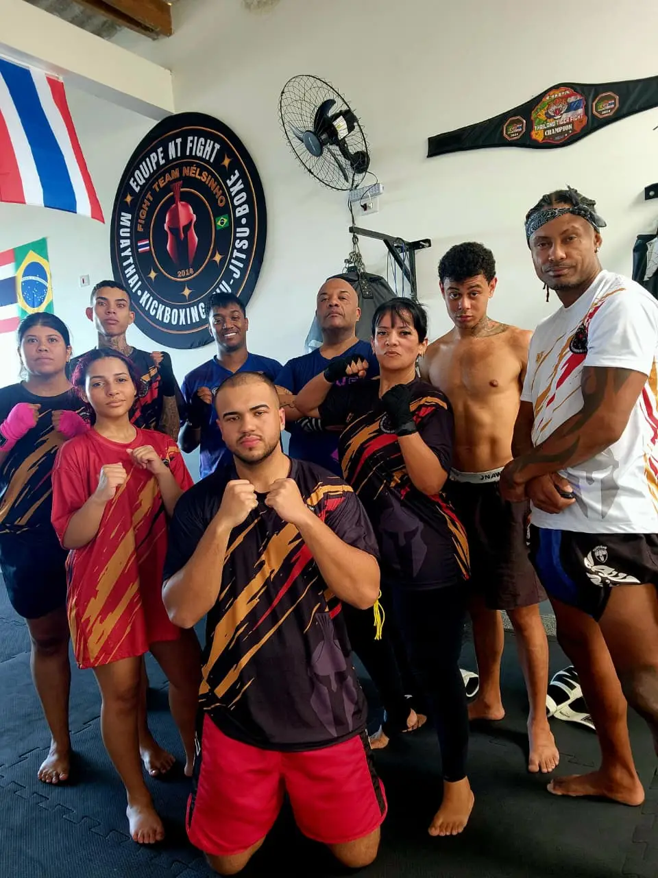 Alunas e alunos da Equipe NT Fight em guarda diante do brasão do CT, com as bandeiras do Brasil e da Tailândia e o cinturão na parede